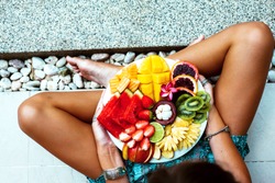 Girl relaxing and eating fruit plate by the hotel pool. Exotic summer diet. Photo of legs with healthy food by the poolside, top view from above. Tropical beach lifestyle.