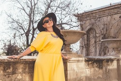 Young stylish woman wearing yellow maxi dress, black hat and sunglasses walking in the city street. Spring fashion outfit, elegant look. Plus size model.
