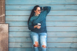 Beautiful woman wearing fall sweater, ripped jeans and glasses drinking take away coffee standing against cafe wall on city street. Casual fashion, elegant everyday look. Plus size model.