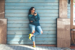 Beautiful woman wearing fall sweater, ripped jeans and colorful shoes drinking take away coffee standing against cafe wall on city street. Casual fashion, elegant everyday look. Plus size model.