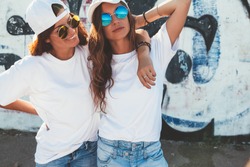 Two models wearing plain white t-shirts and hipster sunglasses posing against street wall. Teen urban clothing style, same look. Mockup for tshirt print store. 
