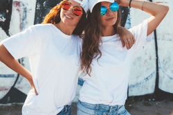 Two models wearing plain white t-shirts and hipster sunglasses posing against street wall. Teen urban clothing style, same look. Mockup for tshirt print store. 