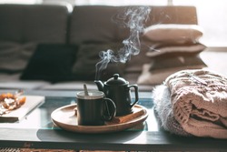 Still life details in home interior of living room. Sweaters and cup of tea with steam on a serving tray on a coffee table. Breakfast over sofa in morning sunlight. Cozy autumn or winter concept.