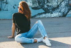 Model wearing plain black t-shirt, boyfriend jeans, sneakers and hipster sunglasses posing against street wall, teen urban clothing style, mockup for tshirt print store