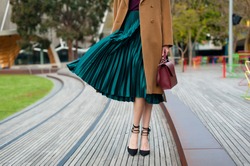 Fashionable young woman wearing green pleated midi skirt, sweater, high heel shoes, beige wool coat and holding burgundy handbag in hand on the city street. Street style.