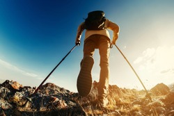 Hiker goes with trekking poles uphill against sunset sky and sun. Hiking concept