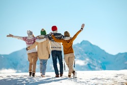 Four happy friends are standing and embracing against snow capped mountains at sunny day. Winter vacations concept