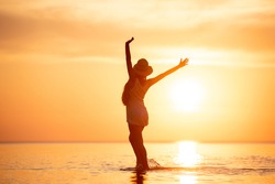 Lady's silhouette stands with raised arms at tranquil sunset beach