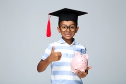 Young African American boy in graduation cap showing thumb up and holding piggybank on grey background