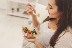 Young pregnant woman eating green salad. Attractive expectant lady sitting on sofa and having fresh snack. Healthy nutrition and pregnancy concept, copy space