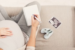 Unrecognizable pregnant woman sitting on sofa with blank notebook, pen, tiny shoes and sonogram and writing names for her baby, making shopping list, top view, copy space