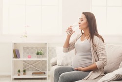 Pregnant woman drinking water sitting on sofa. Young expectant lady have rest at home with glass, copy space. Pregnancy, healthcare, thirst concept