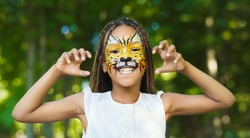Little african-american girl with tiger face painting roaring, making funny grimace outdoors, copy space