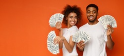 Happy millennial couple posing with lot of money, holding with dollar fans, orange panorama background with empty space