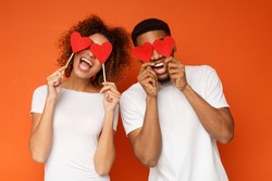 Lovers blinded by their big love.. Young cheerful african-american couple in love holding red hearts over eyes and smiling, orange background