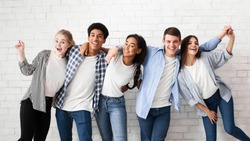 Diverse teens embracing and posing over white brick wall, looking at camera