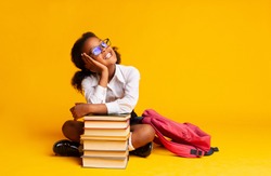 Cheerful African American Schoolgirl Dreaming Sitting At Book Stack Over Yellow Background In Studio. Copy Space