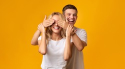 Happy couple playing hide and seek. Young man standing behind his girlfriend and closing her eyes, yellow background with copy space