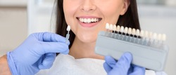 Dentist in blue medical gloves applying sample from tooth enamel scale to happy woman patient teeth to pick up right shade, teeth bleaching procedure, cropped, panorama