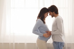 Happy young couple expecting baby standing together against window at home, loving husband tenderly touching belly of his pregnant wife, free space