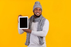 New application or website. Afro man in winter clothes showing blank digital tablet screen, yellow studio background