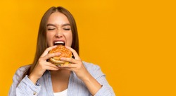 Young Lady Biting Burger Enjoying Junk Food On Cheat Meal Day Standing Over Yellow Background. Panorama, Studio Shot, Copy Space