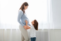 Excited little daughter touching pregnant mom tummy, standing next to window, copy space