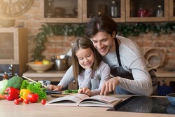 Happy dad and little daughter checking recipe in cookbook at kitchen together while cooking healthy lunch at home, free space