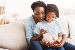 Money Saving Concept. Excited black mom and daughter putting coins into piggy bank, free space