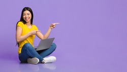 Special Offer. Cheerful asian girl sitting on floor with laptop and pointing at copy space over purple background, panorama