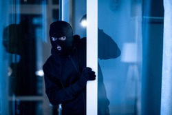 Is Anybody Home. Cautious masked thief looking and peeping into office through window or glass door with torch flashlight