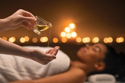 Closeup of female therapist applying massage oil on hands before therapy, young black lady lying on massage table at spa