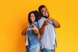 Cheerful African American Couple Standing Back To Back And Pointing At Camera With Fingers Over Yellow Studio Background With Copy Space