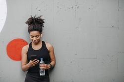 Music and sport. Smiling muscular african american young woman with headphones, smart watch and bottle of water looks at smartphone on wall background, free space