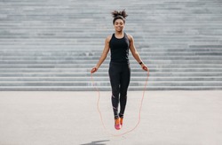 Female athlete does cardio workout. Smiling african american girl in sportswear with fitness tracker jumping rope outdoors, free space