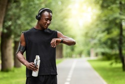 Modern technologies and active lifestyle. African sportsman working out with fitness bracelet, drinking water, blurred park background, copy space