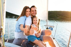 Family Gesturing Thumbs-Up Relaxing On Yacht Posing Sitting On Deck Enjoying Sailing In Sea On Summer Day.