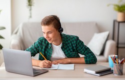 Joyful teen guy writing at notebook and looking at laptop screen, having lesson online, home interior, free space