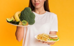 Healthy Vs Unhealthy. Unrecognizable girl choosing between plates with organic fruits and vegetables and junk food, yellow background, cropped