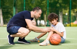 Sports Injury. Handsome PE teacher helping boy with knee trauma after playing basketball