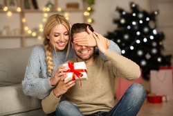 Beautiful woman surprising her husband with Christmas gift at home, closing his eyes
