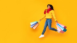 Autumn Sales. African Shopaholic Woman Holding Shopping Bags Running In Mid-Air Wearing Winter Clothes On Yellow Studio Background. Panorama, Copyspace