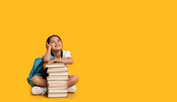 Chinese Elementary School Girl Sitting At Books Stack Posing On Yellow Background. Studio Shot, Free Space For Text