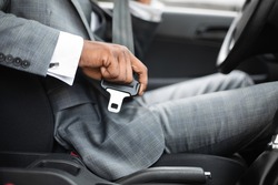Driving safety concept. Unrecognizable black businessman fasten seat belt in his car, ready to go to office. African american man in stylish suit putting on his seatbelt before driving car, cropped