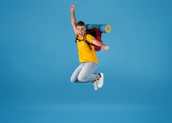 Excited young Caucasian tourist with camping equipment jumping on blue studio background. Full length portrait of funky guy thrilled over his summer holiday camping trip