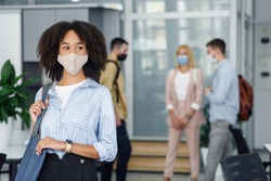 Focus on portrait of modern business african american woman came to work in morning. Colleagues in protective masks with bags in office interior are blurred background