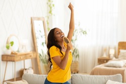 Cheerful black woman with hairbrush as mic singing her favorite song and dancing at home. Millennial African American lady pretending to be famous singer, having fun in living room