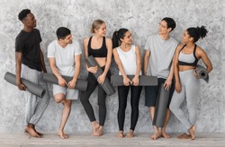 Sporty Lifestyle Concept. Group of athletic multiracial barefoot people resting with yoga mats before training in studio, standing near grey wall, wearing comfortable sportswear, smiling to each other