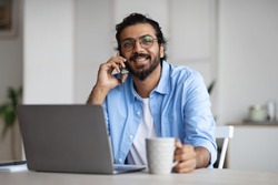 Cheerful Indian Freelancer Guy Drinking Coffee And Talking On Cellphone While Having Break During Work With Laptop Computer At Home Office, Speaking With Friends, Resting At Workplace, Free Space