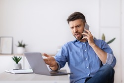 Angry millennial businessman talking on mobile phone and looking at laptop screen working in modern office, having problems with project, annoyed boss speaking with employee by phone, copy space